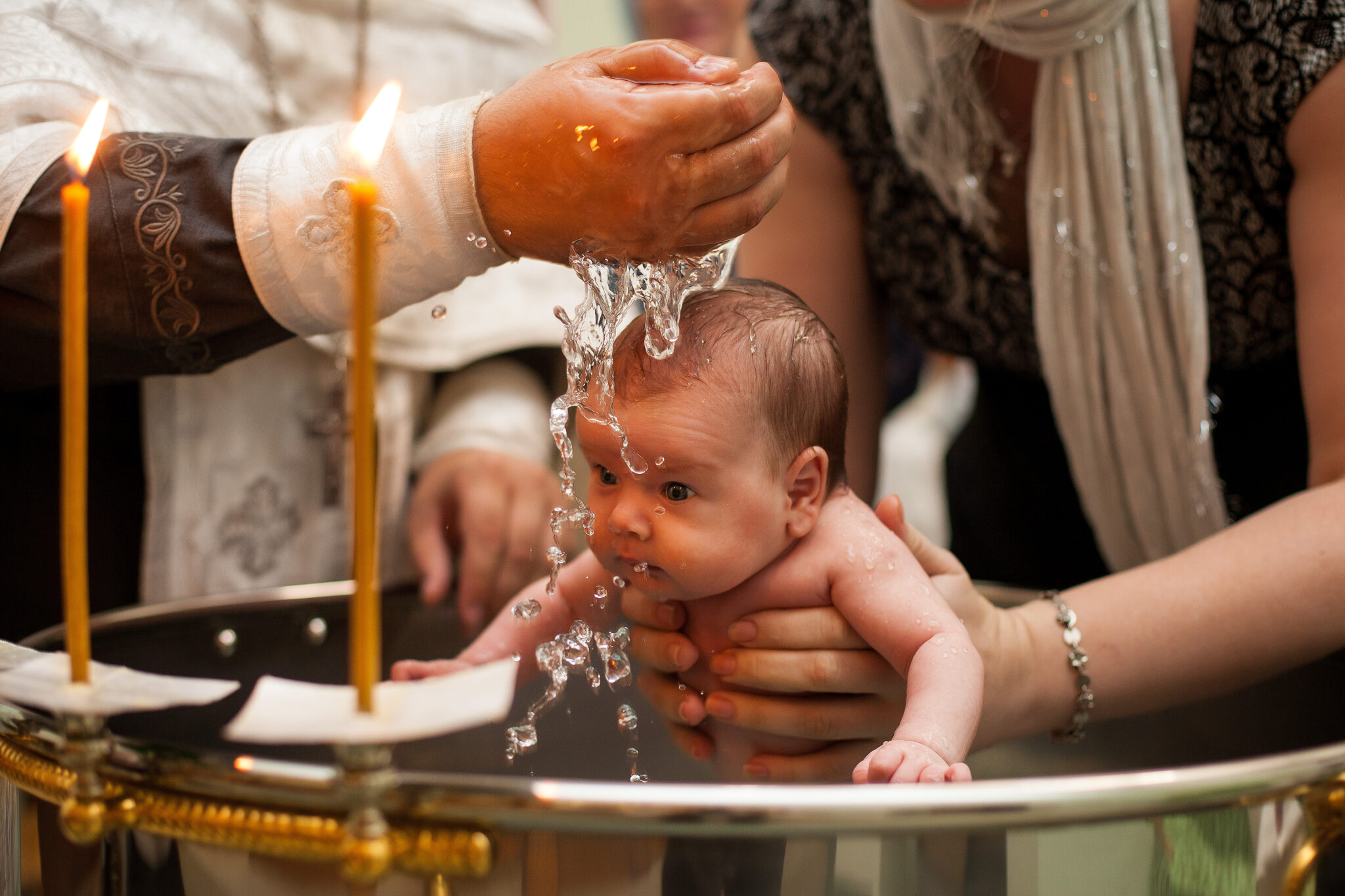 Newborn baby baptism in Holy water. baby holding mother's hands. Infant bathe in water. Baptism in the font. Sacrament of baptism. Child and God. Christening candle Holy water font. The priest baptize